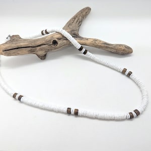 May include: A necklace composed of small, flat, white circular beads strung together, accented with pairs of dark brown cylindrical wooden beads placed at intervals along the strand, resting against a piece of light brown driftwood.