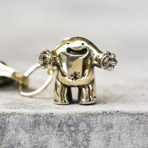 May include: A gold-colored metal keychain featuring a small, cartoon-like figure with outstretched arms and a smiling face. The figure is attached to a metal ring and a leather strap.