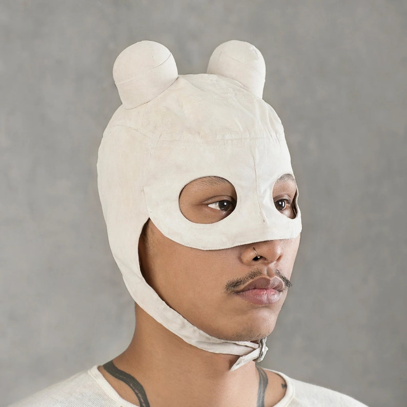 May include: A white fabric mask with eye holes and ear flaps. The mask has a rounded shape and resembles a bear's head. The mask is worn by a person with a nose ring.