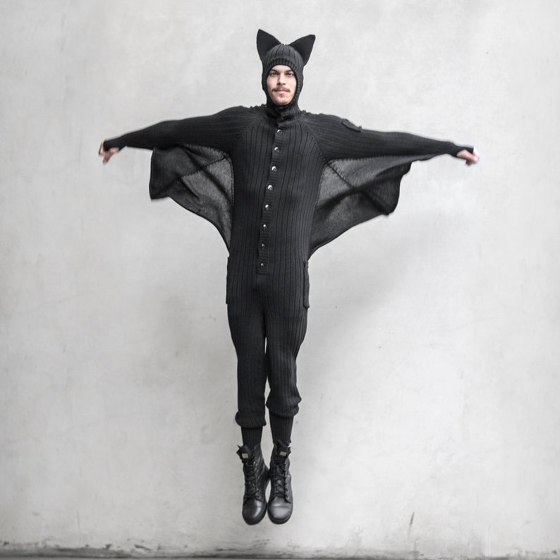 May include: A black knit bat costume with a matching hat and attached wings. The one-piece outfit features a button-up front, pockets, and is paired with black boots. The person is mid-air with arms outstretched, showcasing the costume's design.