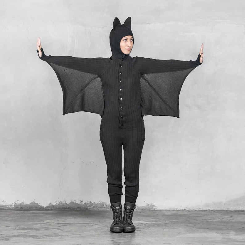 May include: Black knit bat costume with attached wings and hood with bat ears. The one-piece outfit has a button-up front and pockets. The person is wearing black boots. The costume is designed to look like a bat.