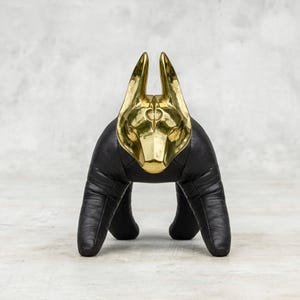 Puede incluir: Escultura decorativa con forma de perro. La cabeza es de metal dorado pulido, con orejas erguidas y detalles faciales. El cuerpo es negro, con apariencia de cuero texturizado, y se apoya en cuatro patas. Diseño moderno y artístico, con contraste de colores.