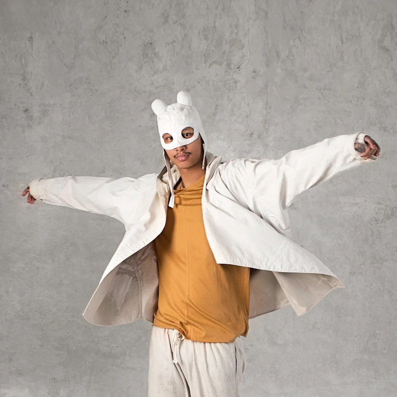 May include: A person wearing a white bunny mask and a white jacket over a yellow shirt and white pants. The person is standing in front of a gray concrete wall.
