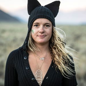 May include: A woman wearing a black knit cat ear beanie and a black button-down sweater. She has long, wavy blonde hair and a gold necklace. A green and white heart-shaped design is on her cheek. The background is a blurred outdoor scene.