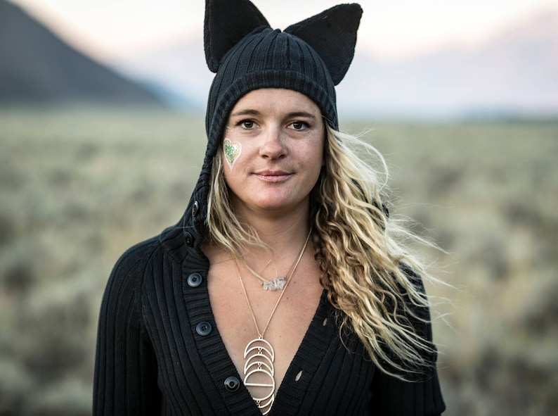 May include: A woman wearing a black knit cat ear beanie and a black button-down sweater. She has long, wavy blonde hair and a gold necklace. A green and white heart-shaped design is on her cheek. The background is a blurred outdoor scene.