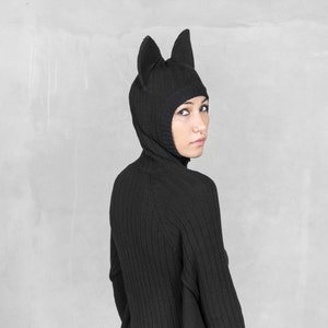 May include: A black knit hood with pointed ears, worn with a matching black ribbed sweater. The hood covers the head and neck, creating a sleek, monochromatic look. The outfit is made of a ribbed knit fabric.
