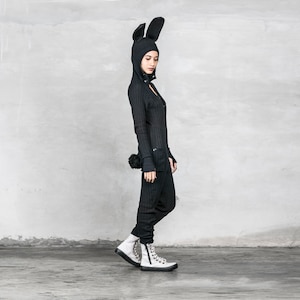 May include: A black bunny costume with a long-sleeved, hooded jumpsuit and a fluffy tail. The costume is worn by a person with black hair and is standing on a gray background.