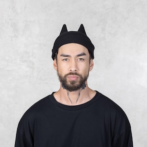 May include: A black knit beanie featuring two pointed, cat-ear-shaped protrusions on the top. The hat is constructed with a ribbed texture and a folded cuff design, providing a snug fit around the head. The material is a solid, matte black color.