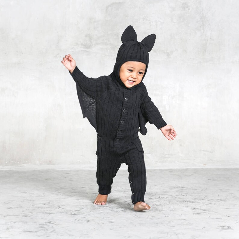 May include: A black knitted baby bat costume. The outfit features a hooded top with bat ears, a button-down front, and attached wings. The matching trousers have button details at the ankles. The baby is barefoot and smiling.