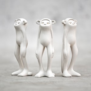 Könnte beinhalten: Drei weiße Keramikfiguren von humanoiden Figuren mit langen Gliedmaßen und großen Köpfen. Die Figuren haben kleine, runde Augen mit goldenen Akzenten.