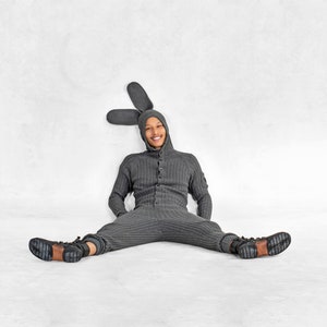 May include: A gray knit jumpsuit with a hood and button-up front, featuring attached bunny ears. The person is wearing black boots with brown soles. The outfit is designed to resemble a rabbit.