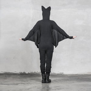 May include: A black knit jumpsuit with attached bat wings and a hood with bat ears. The jumpsuit has long sleeves, a ribbed texture, and pockets on the legs. The person is wearing black boots. The background is a plain gray wall.