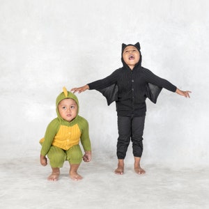 May include: Two children in costumes. One child is wearing a green and yellow dinosaur costume with a hood. The other child is wearing a black bat costume with a hooded jacket and wings. Both children are barefoot.
