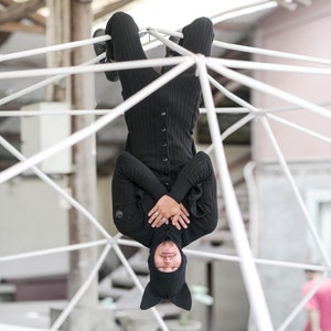 May include: A person wearing a black knit outfit, including a button-down sweater and matching pants, hangs upside down from a white geometric structure. The outfit includes a knit hat with pointed ears. The person's arms are crossed.