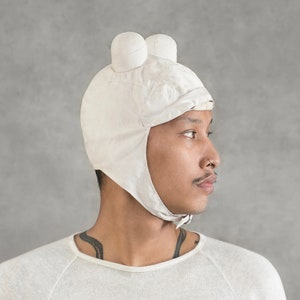 May include: A white cotton hat with two round ear flaps and a chin strap. The hat has a unique design with two round, white ear flaps that resemble ears.