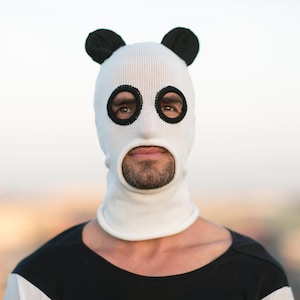 Puede incluir: Un pasamontañas blanco tejido con orejas y aberturas para los ojos negras con forma de panda. El pasamontañas cubre toda la cabeza y el cuello, con una abertura para la boca. La persona lleva una camiseta a rayas blancas y negras.