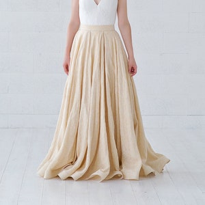 May include: A white V-neck bodice with spaghetti straps is paired with a flowing, light beige skirt. The skirt cascades to the floor. The person is wearing a floral crown. The background is a white brick wall and white floor.