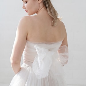 Puede incluir: Una mujer con un vestido de novia blanco sin tirantes con un gran lazo en la espalda. El vestido es de tul y tiene una falda fluida.