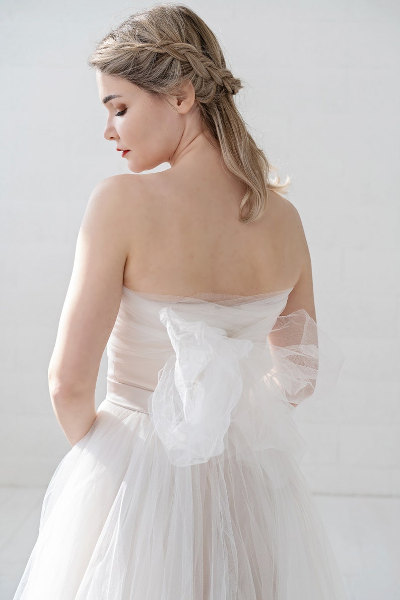 Puede incluir: Una mujer con un vestido de novia blanco sin tirantes con un gran lazo en la espalda. El vestido es de tul y tiene una falda fluida.