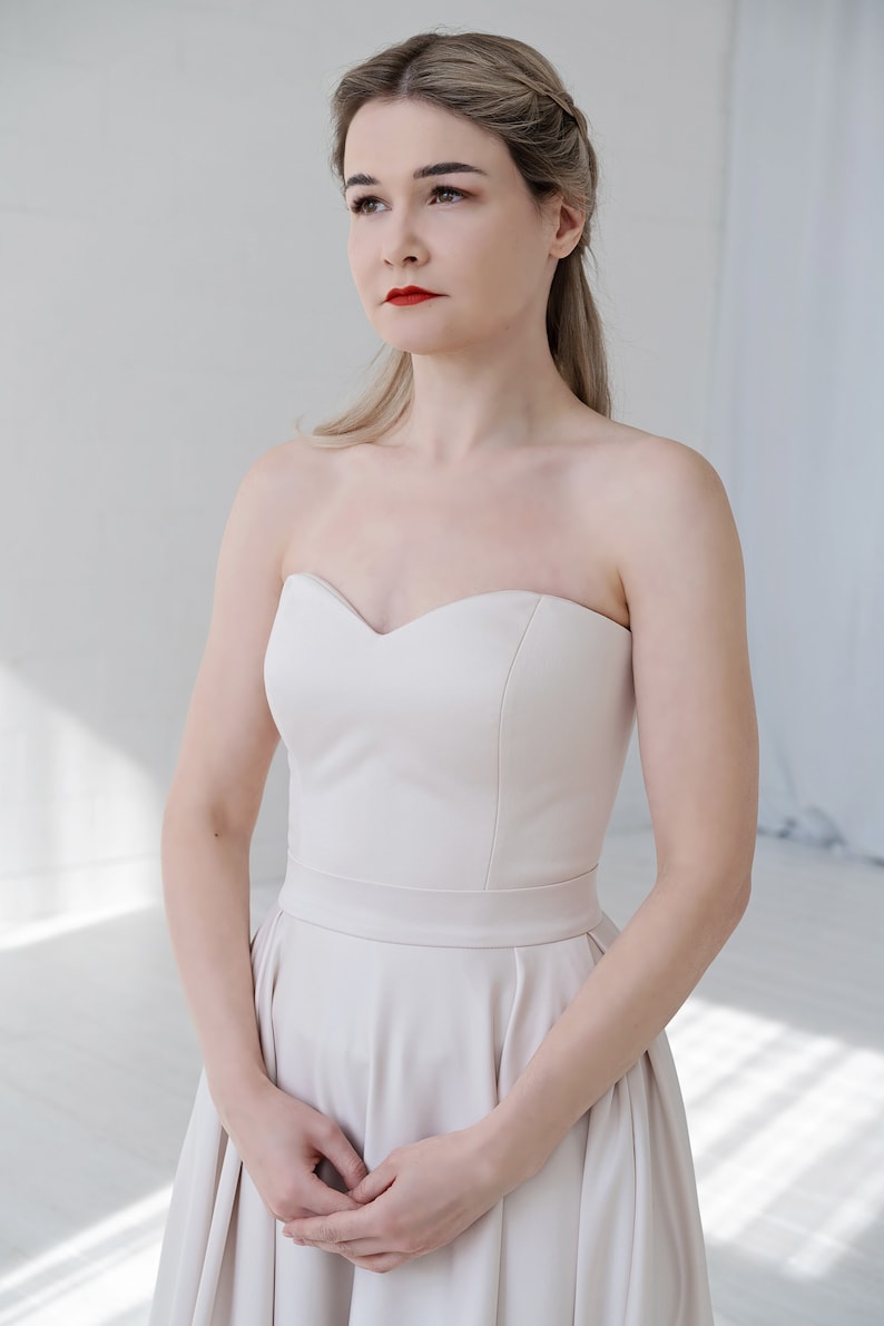 Puede incluir: Una mujer con un vestido de novia de sat&eacute;n marfil sin tirantes con escote de coraz&oacute;n. El vestido tiene un corpi&ntilde;o ajustado y una falda completa.