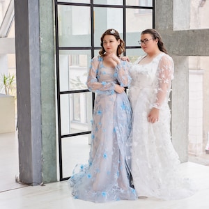 May include: Two women wearing long, flowing dresses. The woman on the left is wearing a light blue dress with floral embroidery. The woman on the right is wearing a white dress with floral embroidery. They are standing in front of a large window.
