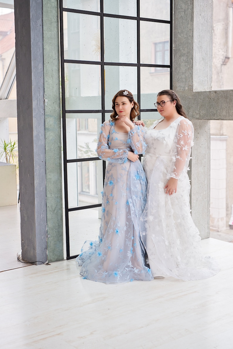 May include: Two women wearing long, flowing dresses. The woman on the left is wearing a light blue dress with floral embroidery. The woman on the right is wearing a white dress with floral embroidery. They are standing in front of a large window.