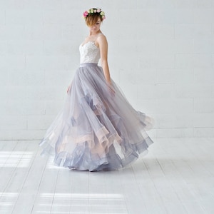 May include: A woman wearing a white lace bodice and a flowing gray tulle skirt with a floral crown. The skirt has multiple layers and is gathered at the waist. The woman is standing on a white wooden floor.