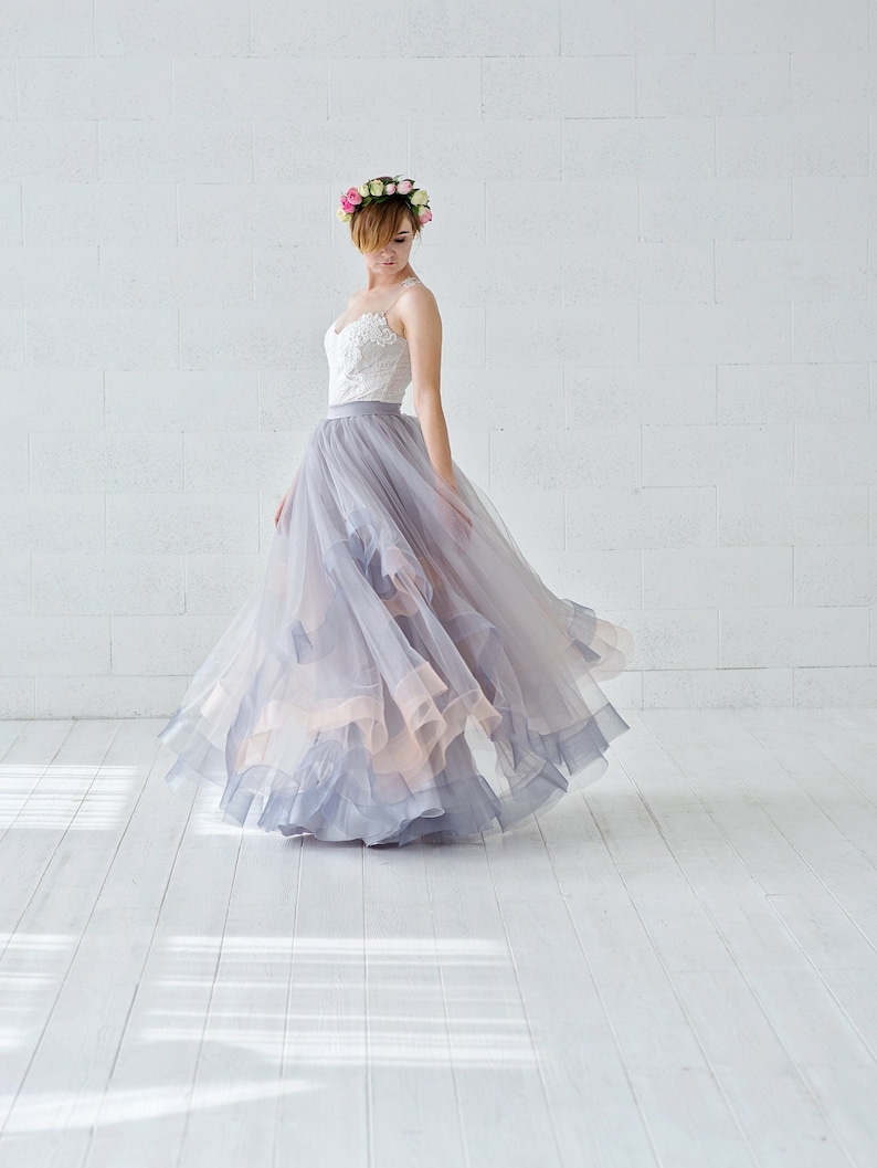 May include: A woman wearing a white lace bodice and a flowing gray tulle skirt with a floral crown. The skirt has multiple layers and is gathered at the waist. The woman is standing on a white wooden floor.
