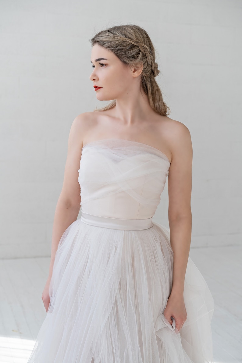 Puede incluir: Una mujer con un vestido de novia blanco sin tirantes con una falda de tul y una banda de sat&eacute;n. Lleva el pelo recogido en una trenza y mira hacia un lado.