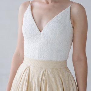 May include: A formal dress featuring a white lace bodice with spaghetti straps and a deep V-neck. The skirt is a light gold color with a subtle pattern and a wide waistband. The dress is suitable for special occasions.