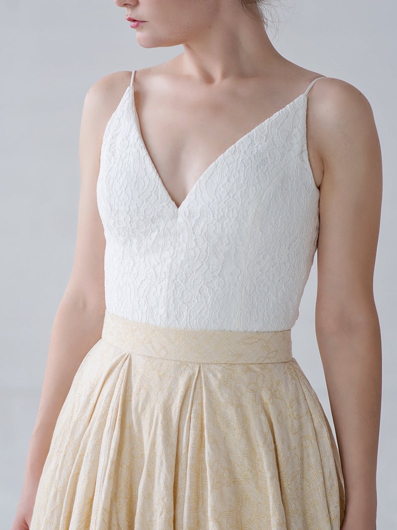 May include: A formal dress featuring a white lace bodice with spaghetti straps and a deep V-neck. The skirt is a light gold color with a subtle pattern and a wide waistband. The dress is suitable for special occasions.