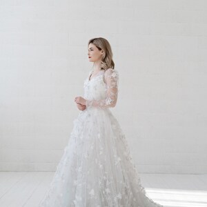 May include: A white wedding dress with long sleeves and a floral design. The dress has a fitted bodice and a full skirt. The dress is made of a delicate fabric and has a romantic feel.