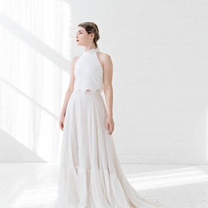 May include: A white halter top with a lace overlay and a white flowing skirt with ruffles. The model is standing in a white room with natural light.