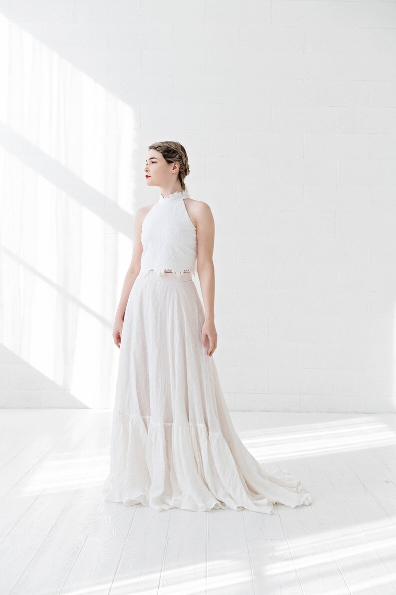May include: A white halter top with a lace overlay and a white flowing skirt with ruffles. The model is standing in a white room with natural light.