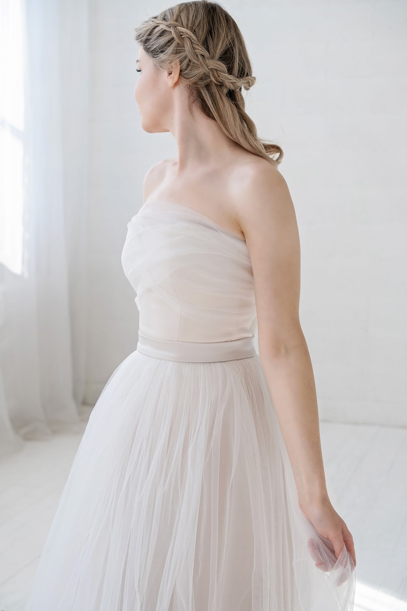 Puede incluir: Un vestido de novia blanco de tul sin tirantes con una banda de sat&eacute;n. El vestido tiene una falda fluida y es perfecto para una novia que quiere un look rom&aacute;ntico y elegante.