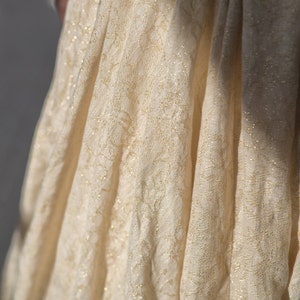 May include: Close-up of a flowing, cream-colored fabric with a delicate floral pattern and subtle gold accents. The fabric is draped, creating soft folds and shadows. The texture appears lightweight and slightly sheer, suggesting a dress or other garment.