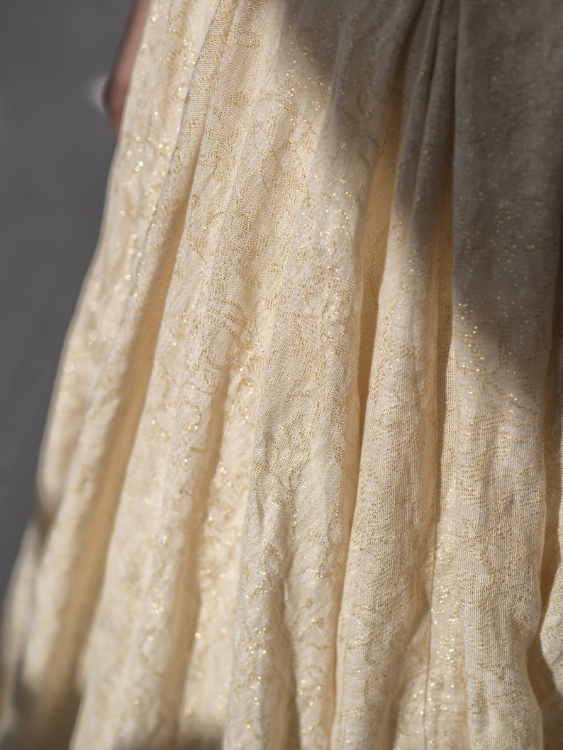 May include: Close-up of a flowing, cream-colored fabric with a delicate floral pattern and subtle gold accents. The fabric is draped, creating soft folds and shadows. The texture appears lightweight and slightly sheer, suggesting a dress or other garment.