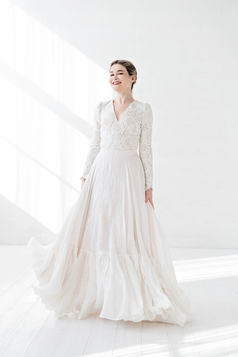 May include: A woman in a white lace top and a flowing white skirt with ruffles. She is smiling and looking to the side.