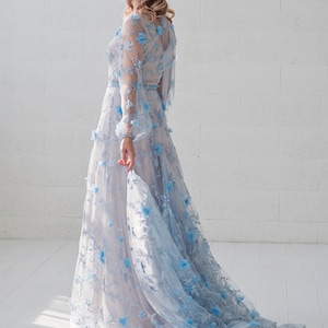 May include: A woman wearing a light blue gown with floral embroidery. The gown has a long train and is made of a sheer fabric.