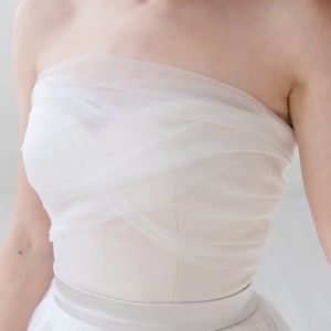 Puede incluir: Un vestido de novia blanco sin tirantes con un corpi&ntilde;o de tul y una falda fluida. El vestido tiene una faja de sat&eacute;n alrededor de la cintura.