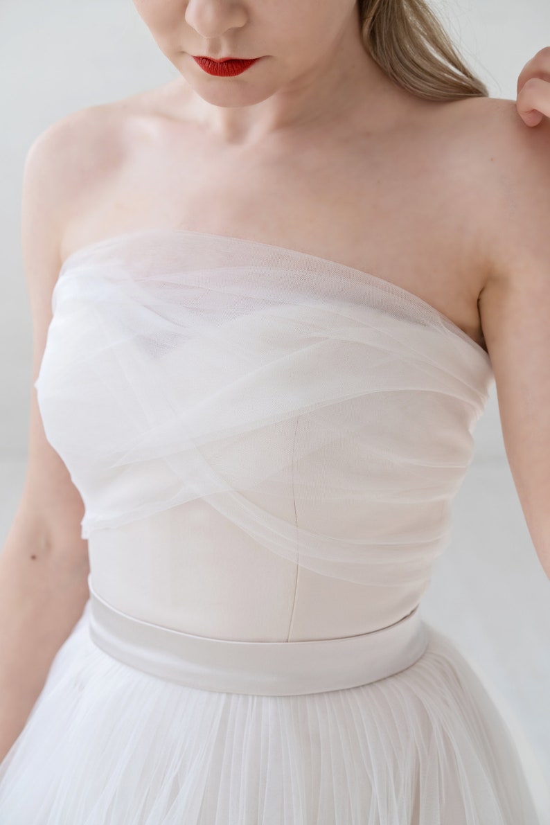 Puede incluir: Un vestido de novia blanco sin tirantes con un corpi&ntilde;o de tul y una falda fluida. El vestido tiene una faja de sat&eacute;n alrededor de la cintura.
