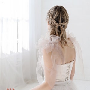 Puede incluir: Una mujer con un vestido de novia de tul blanco con un corpi&ntilde;o de sat&eacute;n y una delicada superposici&oacute;n de tul rosa. Su cabello est&aacute; peinado en una corona trenzada.