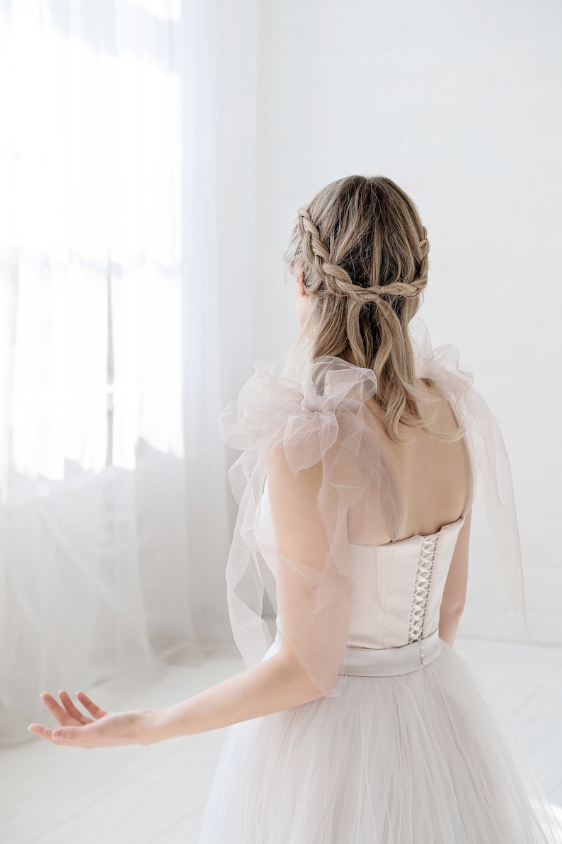 Puede incluir: Una mujer con un vestido de novia de tul blanco con un corpi&ntilde;o de sat&eacute;n y una delicada superposici&oacute;n de tul rosa. Su cabello est&aacute; peinado en una corona trenzada.