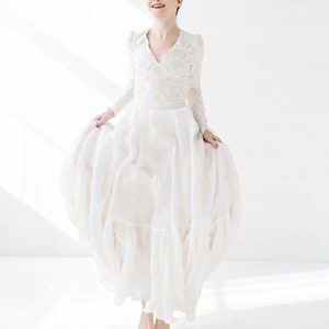 May include: A woman in a white lace top and a flowing white skirt jumps in the air, smiling at the camera. The background is a white wall with natural light.