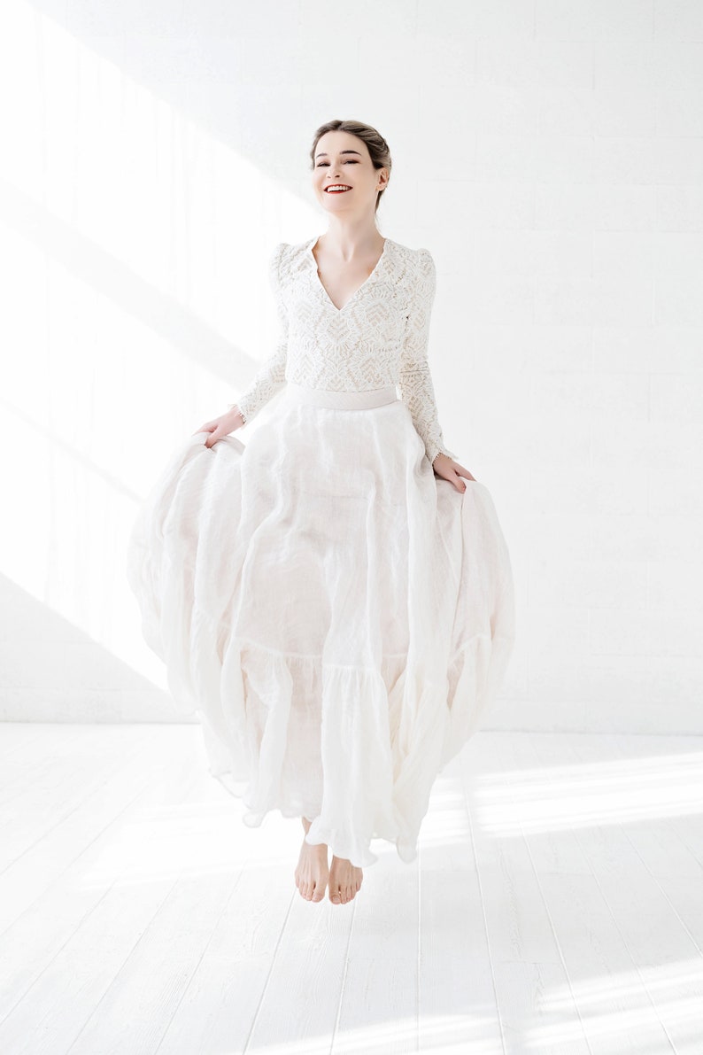 May include: A woman in a white lace top and a flowing white skirt jumps in the air, smiling at the camera. The background is a white wall with natural light.