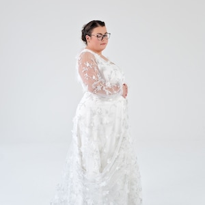 May include: A white lace wedding dress with long sleeves and a flowing skirt. The dress has a delicate floral pattern and is worn by a person with dark hair and glasses.