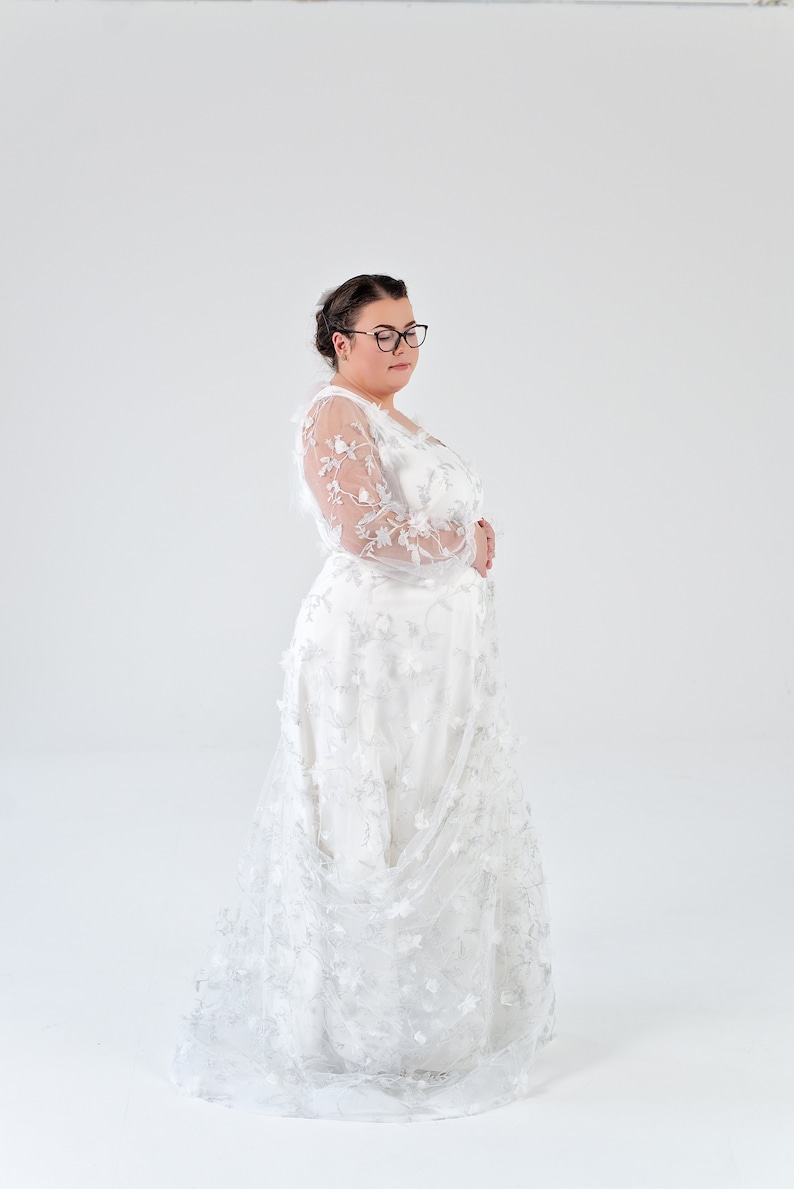 May include: A white lace wedding dress with long sleeves and a flowing skirt. The dress has a delicate floral pattern and is worn by a person with dark hair and glasses.