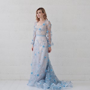 May include: A woman wearing a light blue, long-sleeved gown with floral embroidery. The gown has a fitted bodice and a flowing skirt.