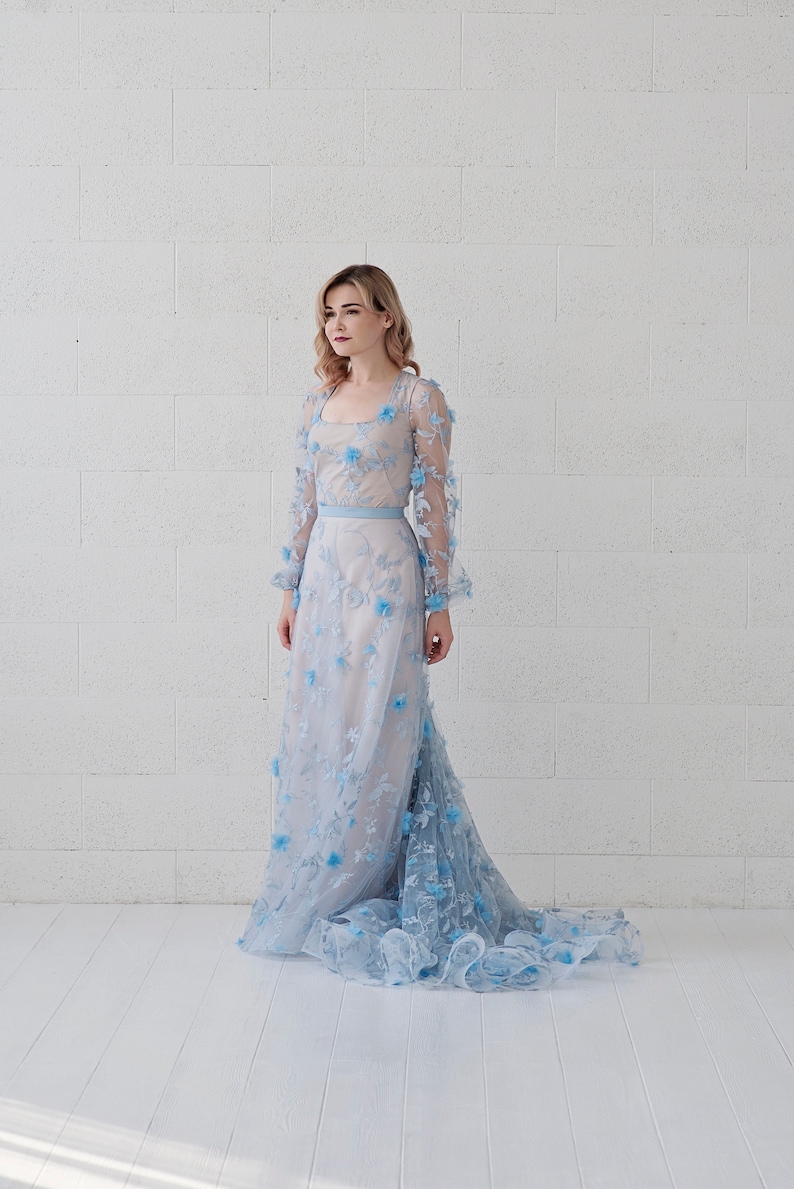 May include: A woman wearing a light blue, long-sleeved gown with floral embroidery. The gown has a fitted bodice and a flowing skirt.
