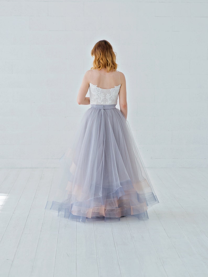 May include: A woman wearing a white lace top and a layered tulle skirt in shades of gray and peach. The skirt is full and flowing, and the top is fitted.